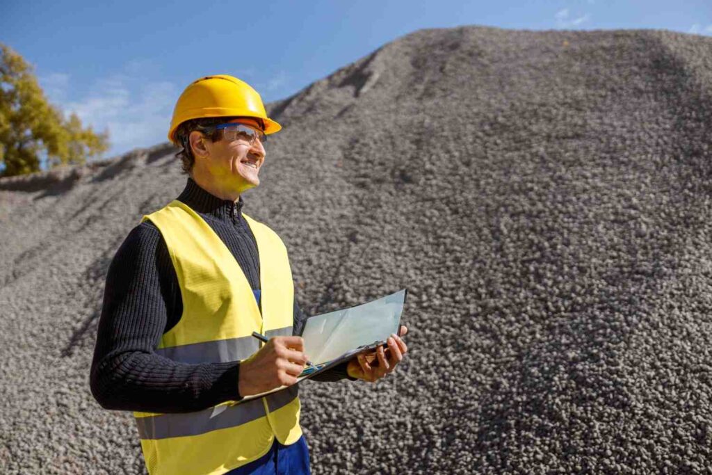 Engenheiro de mineração com capacete e colete de segurança registrando dados em frente a pilha de brita.