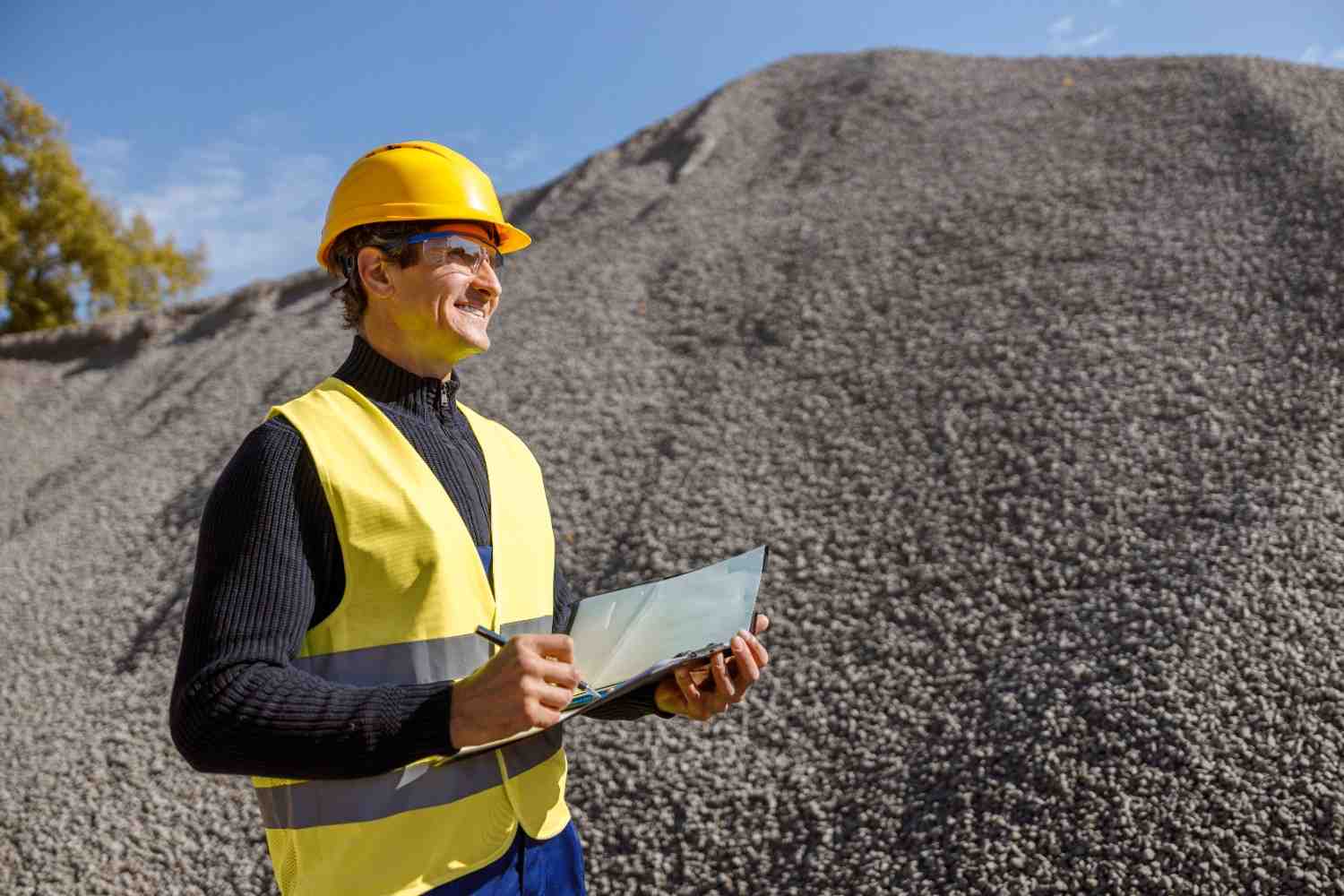 Engenheiro de mineração com capacete e colete de segurança registrando dados em frente a pilha de brita.